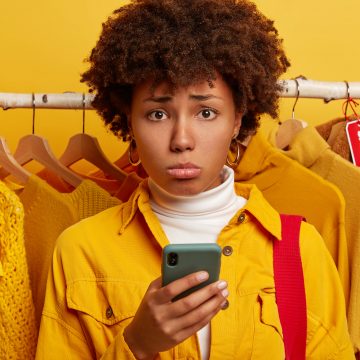 Dejected Afro woman uses smartphone for online shopping, unhappy cannot find schedule of sales, stands against yellow clothes in hangers with price cut, has no suitable size. Shopping season